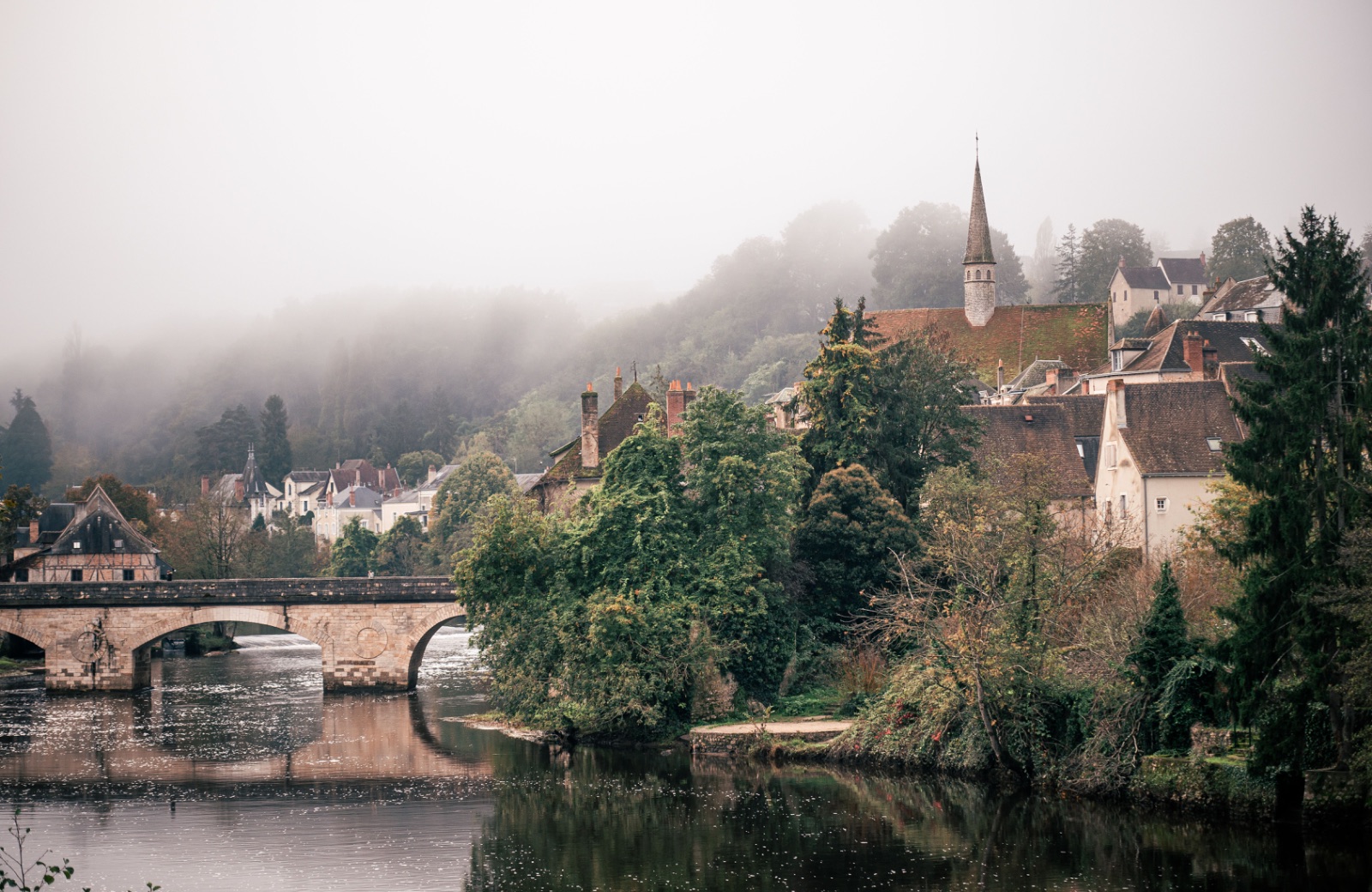 Misty French riverside village with stone arched bridge, church spire and stone houses above wooded banks